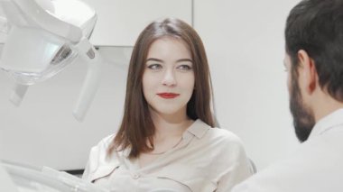 Modern bir dişçi kliniğindeki aynada ışıldayan gülüşüne hayran olan büyüleyici genç bir kadın kendinden emin bir şekilde ışıldıyor. Parlak, temiz ortam dişlerini kontrol ettikten sonra neşesini arttırır. Ağız sağlığının önemini gösterir.