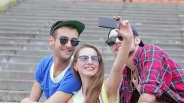 Canlı bir üçlü arkadaş, güneşin tadını çıkarıyor, merdivenlerde eğlenceli bir selfie çekiyor, mutluluk ve dostluk yayıyor. Şık güneş gözlükleri ve canlı kıyafetleri kaygısız bir yaz atmosferini yansıtıyor ve bu anı unutulmaz kılıyor.