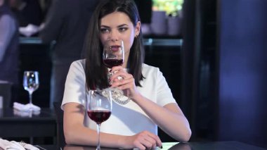 Şık bir esmer kadın, modern bir restoran ortamında bir bardak zengin kırmızı şaraptan zevk alır. Elinde beyaz bir akıllı telefonla, o anı yakalar, rahatlamayla teknolojiyi harmanlayarak canlı bir atmosferde.