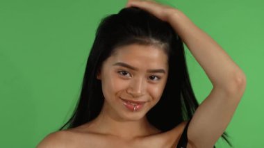Büyüleyici bir stüdyo portresi, parlak yeşil renkli arka plan üzerinde uzun, dalgalı saçlarını şakacı bir şekilde kıvırırken neşe saçan dudak halkalı, çarpıcı bir Asyalı genç kadını gösteriyor. Kendine güveni ve doğal güzelliği davetkar bir güzellik yaratır.