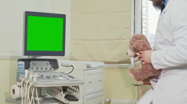 Beyaz önlük giymiş neşeli bir çocuk doktoru, canlı yeşil ekranlı ultrason tarayıcısının yanında otururken yumuşak bir oyuncak ayı tutuyor. Bu iç açıcı sahne, çocuk dostu sağlık hizmetlerinin özünü yansıtıyor.
