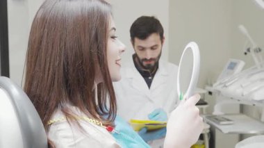 Neşeli genç bir kadın dişlerini kontrol ettikten sonra kameraya doğru ışıldıyor. Parlak bir klinikte, profesyonel bir dişçi tedavisini dikkatle incelerken aynada gülümsemesini takdir eder.
