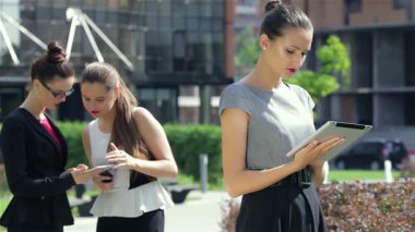 Tarz sahibi gri bluz ve siyah pantolon giyen dengeli genç bir iş kadını, elinde bir tabletle güvenle ayakta dururken, bayan meslektaşları da yakınlarda ortak bir tartışma içinde. Bu dinamik sahne takım çalışmasının özünü yakalar ve...