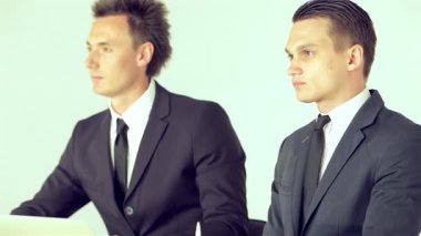 İki keskin giyimli iş adamı, biri parlak siyah takım elbiseli, diğeri şık gri takım elbiseli, bir iş konferansında bir konuşmacıyı dikkatle dinliyorlar, ifadeleri bağlılığı ve profesyonelliği yansıtıyor. Konuşmacı bitirdiğinde, bir araya gelirler.