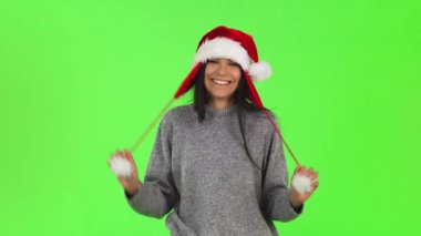 Neşeli genç bir kadın, kaprisli bir Noel Baba şapkası takıyor, neşeli başlığını ayarlarken parlak yeşil renkli arka planlı canlı bir stüdyoda gülüyor. Bu canlı sahne, tatil ruhunun, kutlamanın ve