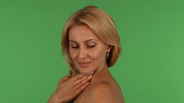 Çekici, olgun bir kadın, dalgalı sarı saçlarıyla kameraya baştan çıkarıcı bir şekilde gülümsüyor, eli, canlı yeşil renkli bir arka plana nazikçe tenini okşuyor. Bu büyüleyici stüdyo portresi özgüven ve şehvet yayıyor.