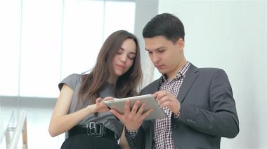 Profesyonel kıyafetli, keyifli genç bir çift şık bir tablet üzerinde etkileyici iş sonuçlarını değerlendiriyor. Heyecan ve takım çalışması yayıyorlar. İfadeleri zaferi ve işbirliğini yansıtıyor, girişimciliğin canlı enerjisini sergiliyor.