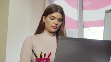 Görünüşte stresli ve yorgun, çekici bir kız öğrenci, masasında oturur, dizüstü bilgisayarında çalışırken şakaklarını ovuşturur. Yumuşak ışıklandırma, mezuniyet sınavlarına hazırlanma karmaşasının ortasında kararlılığını vurguluyor.