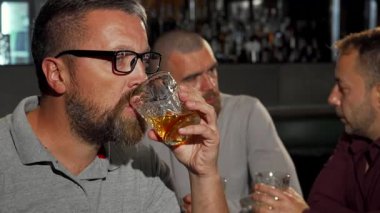 Sakallı olgun bir adam kameraya ışıldıyor, bir bardak zengin viskinin tadını çıkarıyor, arkadaşlarıyla canlı bir bar atmosferinde. Sahne, yoldaşlığın ve kutlamaların özünü yakalar. Arka planda kahkahalar ve bardaklar yankılanır.