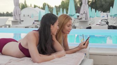 Biri uzun siyah saçlı, diğeri dalgalı sarı bukleli iki güzel kadın, parlak bir yüzme havuzunda dinlenirken akıllı telefonlarına sarılıyorlar. Bu canlı sahne yaz tatilinin özünü yansıtıyor, dostluk gösterisi ve...