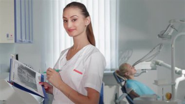 Uzun kahverengi saçlı kendine güvenen bir dişçi, parlak bir diş kliniğinde günlük ve kalem tutarken sıcak bir şekilde gülümsüyor. Arka planda, sakin bir hasta dişçi koltuğuna yaslanıyor, profesyonel ve davetkar bir atmosfer sergiliyor.