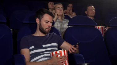 Çizgili patlamış mısır kovası tutan genç bir adam, bir telefon görüşmesine daldı ve yakınlardaki izleyicilerin film deneyimini bozdu. İzleyicilerin rahatsız edici ifadeleri teknoloji ve eğlence arasındaki gerilimi vurguluyor.