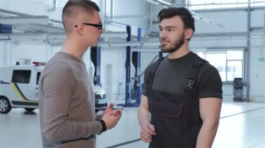 Odaklanmış esmer bir tamirci, modern bir atölyede araba tamirini tartışan gözlüklü genç bir müşteriyle takılır. İki adam el sıkışıyor, profesyonellik gösteriyor ve otomotiv hizmetlerine güveniyorlar.