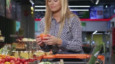 Neşeli sarışın bir kadın, canlı bir süpermarkette domates kokusunun tadını çıkararak alışveriş arabasının üzerine yaslanıyor. Işıl ışıl gülüşü ve şık elbisesi atmosfere renk katıyor. Müşterileri sağlıklı seçimleri kucaklamaya davet ediyor.