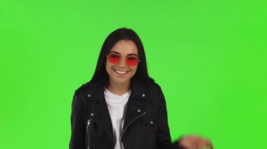 Koyu renk saçlı, şık güneş gözlüklü, heyecan dolu neşeli bir genç kadın elinde parlak yeşil renkli bir arka plana karşı birkaç alışveriş torbası tutuyor. Baş parmak hareketi güven ve neşe saçıyor. Heyecanı mükemmel bir şekilde yakalıyor.