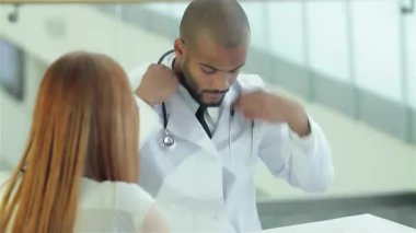 Gıcır gıcır beyaz önlük giyen kendini işine adamış bir doktor, şık ve çağdaş bir klinikte müşterisiyle sağlık sorunlarını dikkatlice tartışıyor. Atmosfer profesyonelce ama davetkar, sağlık hizmetlerinde iletişimin önemini vurguluyor.