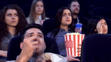 Bir grup genç film izleyicisi loş bir sinemada oturur, dokunaklı bir film izlerken derin duyguları yansıtan yüz ifadeleri. Kıvırcık saçlı beyaz kız ağzını şokla kapatırken, diğerleri dikkatlice bakıyor, elinde patlamış mısır, tamamen daldırılmış.