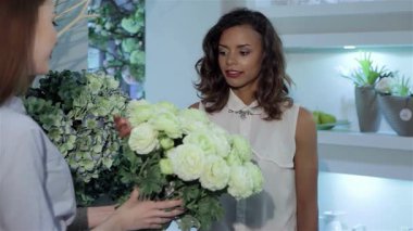 Kıvırcık saçlı genç, güzel bir kadın canlı bir çiçekçide el değmemiş beyaz gül buketini inceliyor. Dost bir çiçekçi zarif düzenlemeyi sunarken, o yumuşak yaprakları incelerken sahne onun narin dokunuşunu yakalar.
