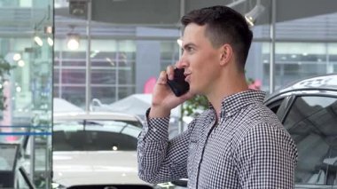 Neşeli genç bir adam, şık bir galeride akıllı telefonuyla sohbet ederken bir elinde arabanın anahtarlarını tutarken heyecanla ışıldıyor. Parlak araçlarla çevrili yeni araba alımının heyecan verici haberini paylaşırken coşkusu yayılıyor.