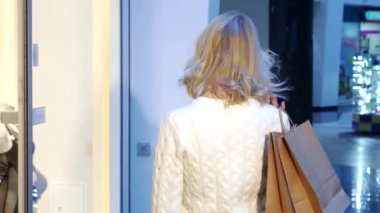 Şık bir iç çamaşırı butiğinin yanında alışveriş torbaları taşırken parlak bir tebessümle, dalgalı sarı saçlı şık bir beyaz kadın arkasına bakıyor. Gündelik ama zarif kıyafeti alışveriş merkezinin hareketli atmosferini mükemmel bir şekilde tamamlıyor.