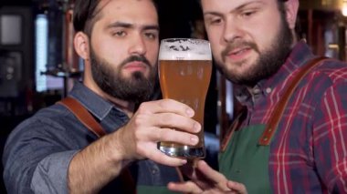 İki yetenekli bira imalatçısı, biri kot gömlekli, diğeri ekose gömlekli, bir bardak altın zanaat birasını yakından inceliyor, bira yapımında uzmanlıklarını sergiliyorlar. Arka planda pırıl pırıl çelik ekipmanlarıyla bira fabrikasının sıcak atmosferi,