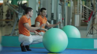 Turuncu tişörtlü odaklanmış genç bir adam dinamik bir antrenman yapar ve çekirdek gücünü ve dengesini arttırır. Spor salonları, aynadaki yansımalarıyla hareketli atmosfer, onun fitness ve atletizme olan bağlılığını vurguluyor.
