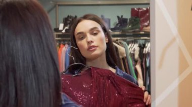 Güzel genç bir kadın şık bir soyunma odasında önünde parlak bir elbise tutarak kameraya ışıldıyor. Havalı kıyafetlerle çevrili, neşe ve güven saçıyor, mükemmel kıyafet için alışverişin heyecanını somutlaştırıyor.