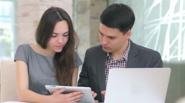 Genç, çekici bir iş kadını ve işadamı, modern bir ofis masasında otururken düzgün bir tabletteki verileri analiz ederek derin bir tartışmaya girişiyor. Odaklanmış ifadeleri ve işbirlikçi ruhları takım çalışmasının özünü vurgular.