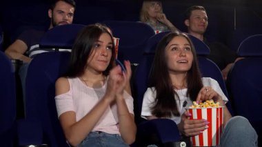 İki neşeli bayan arkadaş, biri uzun siyah saçlı, diğeri açık kahverengi saçlı, sinemanın tadını çıkarıyorlar, kahkahaları ve patlamış mısırları samimi bir tiyatroda paylaşıyorlar. Multiplex 'in canlı atmosferi onların dostluğunu arttırır.