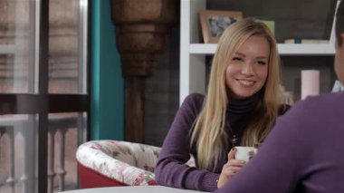 Uzun sarı saçlı neşeli genç bir kadın, beyaz bardaktan çay yudumlarken erkek arkadaşının şakasına yürekten güler. Çiçek dekoru ve hafif ışıklandırmayla süslenmiş davetkar kafe atmosferi, bu şekilde neşeli bağlantılarını güçlendirir.