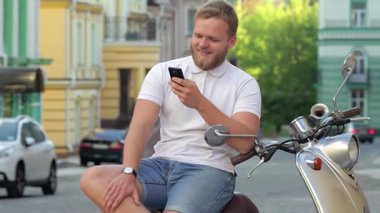 Çekici sakallı genç bir adam, sıcak yaz güneşinin tadını çıkarırken akıllı telefonuna dalmış, motosikletine binmiş öylesine oturuyor. Rahatlamış tavrı ve neşeli gülüşü tasasız bir yaşam tarzını yansıtıyor. Şehir macerasının özünü mükemmel bir şekilde yansıtıyor.