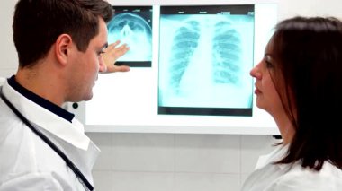 Odaklanmış genç bir erkek doktor, her ikisi de kıtır beyaz laboratuvar önlükleri giymiş olan dikkatli bayan meslektaşına x-ray bulgularını tutkuyla açıklar. Yakın plan çekimleri, parlak görünümde gösterilen x-ray görüntülerini analiz ederken onların meşgul yüz ifadelerini yakalar.