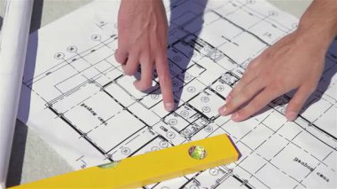 Yakından bakıldığında, erkek parmaklar bina inşaatı planının karmaşık çizgilerini ustalıkla takip ederken, bir kat yakınlarda duruyor. İki adam işbirliği yapıyor, ellerini dinamik bir tartışmaya sokuyorlar, takım çalışması ve hassasiyet sergiliyorlar.