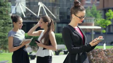 Başarılı genç bir iş kadını dışarıda duruyor, tabletine odaklanmış, kendine güven ve neşe saçıyor. Arka planda, iki meslektaş sohbete dalıp, işbirliği ve profesyonellikten oluşan dinamik bir atmosfer yaratıyorlar.