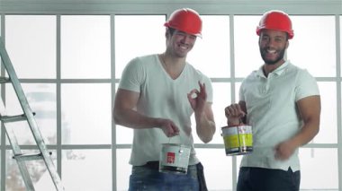 Parlak, boş bir dairede, turuncu kasklı iki neşeli işçi boya kutularını tutarken gururla başparmaklarını kaldırıyorlar. Gülüşleri, inşaat ve yenileme ve vitrinlerle mekanı dönüştürmeye hazırlanırken pozitiflik saçıyor.