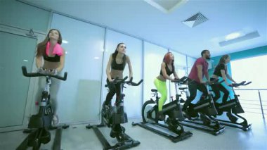 Canlı bir sahne, beş enerjik arkadaşın durağan bisikletlere güçlü bir şekilde pedal çevirmesiyle, dikkatleri modern bir spor salonuna yöneldi. Şık spor kıyafetleri içinde, azim ve dostluk yayıyorlar, fitness ruhunu somutlaştırıyorlar.