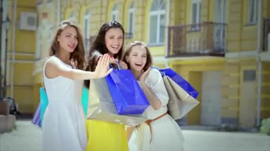 Üç neşeli kız, her biri renkli alışveriş torbaları taşıyor, neşeyle el sallıyor ve hareketli bir alışveriş bölgesinin dışındaki arkadaşlarına gülümsüyorlar. Kaygısız kahkahaları ve şık kıyafetleri mutluluk saçıyor, arkadaşlığın ve eğlencenin özünü yakalıyor.