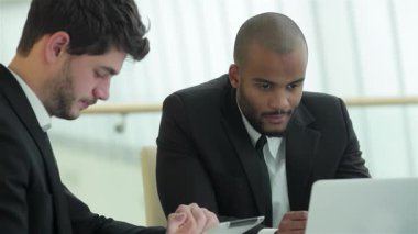 İki başarılı iş adamı, biri siyah saçlı, diğeri de kafası tıraşlı, gösterişli bir ofis masasında verimli bir toplantı yapıyorlar. Özgüvenli ifadeleri ve profesyonel kıyafetleri takım çalışmasının dinamik atmosferini yansıtıyor.