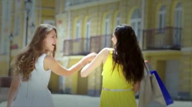 Biri dalgalı beyaz elbiseli, diğeri canlı sarı elbiseli iki şık kız, el ele yürüyorlar, hareketli alışveriş bölgesini keşfederken neşe saçıyorlar. Neşeli gülümsemeleri ve renkli alışveriş torbaları heyecanın vücut bulmuş halidir.