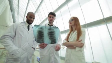 Parlak, modern bir klinikte, üç kendine güvenen doktor, iki erkek ve bir kadın, uzmanlıklarını göstermek için sağlıklı akciğerlerin röntgenini incelerler. Atmosfer, imajın etkilerini tartıştıkları için profesyonellik ve takım çalışması ile dolu.