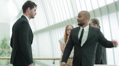 İki başarılı iş adamı dostane bir tokalaşma içindeler. Parlak, çağdaş bir ofis ortamında güven ve dostluk yayıyorlar. Gülüşleri ortak bir zaferi yansıtırken, arka plandaki meslektaşları da canlı atmosfere katkıda bulunuyor.