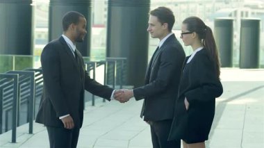Canlı bir sahne, resmi giyinmiş gülümseyen bir grup meslektaşla çevrili iki iş adamı arasındaki dinamik tokalaşmayı yakalıyor. Modern şehir sokaklarındaki bu ilgi çekici an işbirliğini ve yeni şehrin heyecanını simgeliyor.