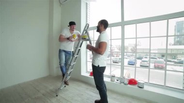 İki neşeli işçi yeni yenilenmiş bir dairede atıştırmalık ve kahkaha paylaşıyorlar. Bir adam, merdivene tünemiş, ellerini meslektaşına boyuyor, doğal ışıkla dolu, parlak, havadar bir alanda takım çalışması ve dostluk sergiliyor.