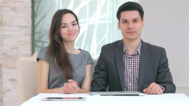 Çekici genç bir çift, şık takım elbiseli bir adam ve şık elbiseli bir kadın, parlak, modern bir çalışma alanında birlikte oturuyorlar, kameraya güvenle gülümsüyorlar. Çekici ifadeleri, önlerindeki parlak dizüstü bilgisayarı ve tableti yansıtıyor.