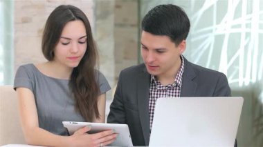 Odaklanmış genç bir iş adamı ve kadın, modern ofis ortamlarıyla çevrili şık bir tablet üzerindeki verileri analiz ederken canlı bir tartışmaya girişiyor. İfadeleri kararlılığı ve takım çalışmasını yansıtıyor.