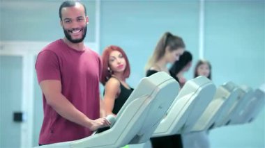 Kendinden emin bir sporcu, canlı bir spor salonu ortamında odaklanmış kişilerle çevrili bir koşu bandında koşarken kameraya gülümsüyor. Sahne fitness 'in dinamik enerjisini yakalıyor, farklı sporcuları şık spor kıyafetleriyle sergiliyor,