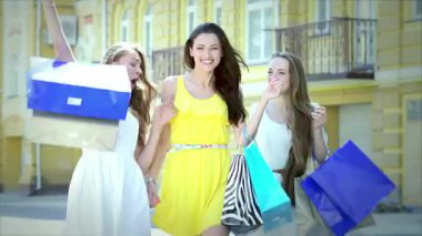 Canlı kıyafetler giymiş üç neşeli kız, güneşli bir caddede geziyorlar, renkli alışveriş çantalarını gösteriyorlar. Gülüşleri ve gülüşleri arkadaşlığın ve kaygısız alışveriş maceralarının özünü yakalar, her anı hissettirir.