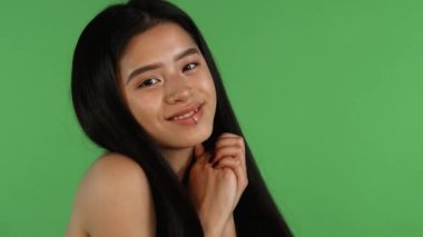 Neşeli bir Asyalı kadının büyüleyici portresi. Uzun, dalgalı saçları var. Ellerini şakaya şakaya tutarak gülümsüyor. Canlı yeşil renkli arka plan onun neşeli tavrını güçlendiriyor, gençliğin ve pozitifliğin özünü somutlaştırıyor.