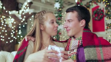 Sıcak ve samimi bir sahne, hoş bir battaniyeye sarılı bir çifti, güzel paketlenmiş hediyelerini, şenlikli bir odada paylaşırken yakalar. Süslü süveter ve eşarplar giymiş genç adam ve kadın, şefkatli bir gülümseme paylaşıyorlar.