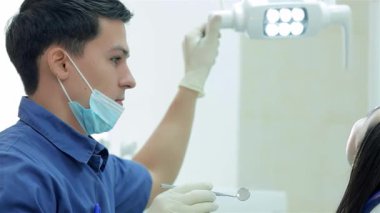 Mavi üniforma ve koruyucu maske takan odaklanmış bir dişçi, tepedeki lambayı optimal görüş için ayarlarken diş aletleri tutuyor. Hasta sandalyeye yaslanıyor, tam bir diş muayenesi için hazır, bir profesyoneli gösteriyor.